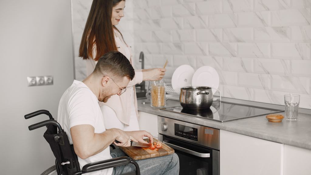 A man in a wheelchair chopping vegetables at a kitchen worktop while a carer stands nearby.