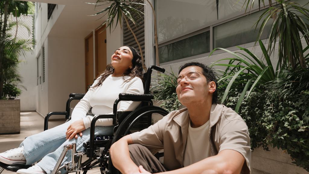 A woman using a wheelchair and a man beside her outdoors, relaxed and talking in the sun.