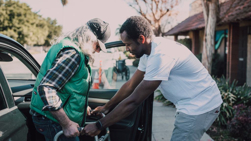 A carer supporting an older man as he gets out of a car, with a wheelchair ready beside them.