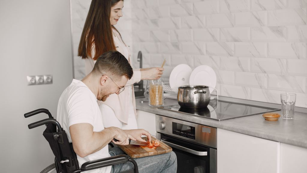 A young man in a wheelchair preparing food at his kitchen worktop while a carer stands alongside him.