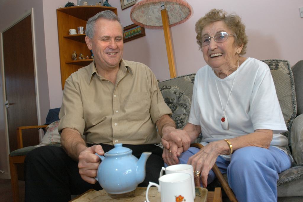 An older couple sitting together at home with a pot of tea, talking and laughing.