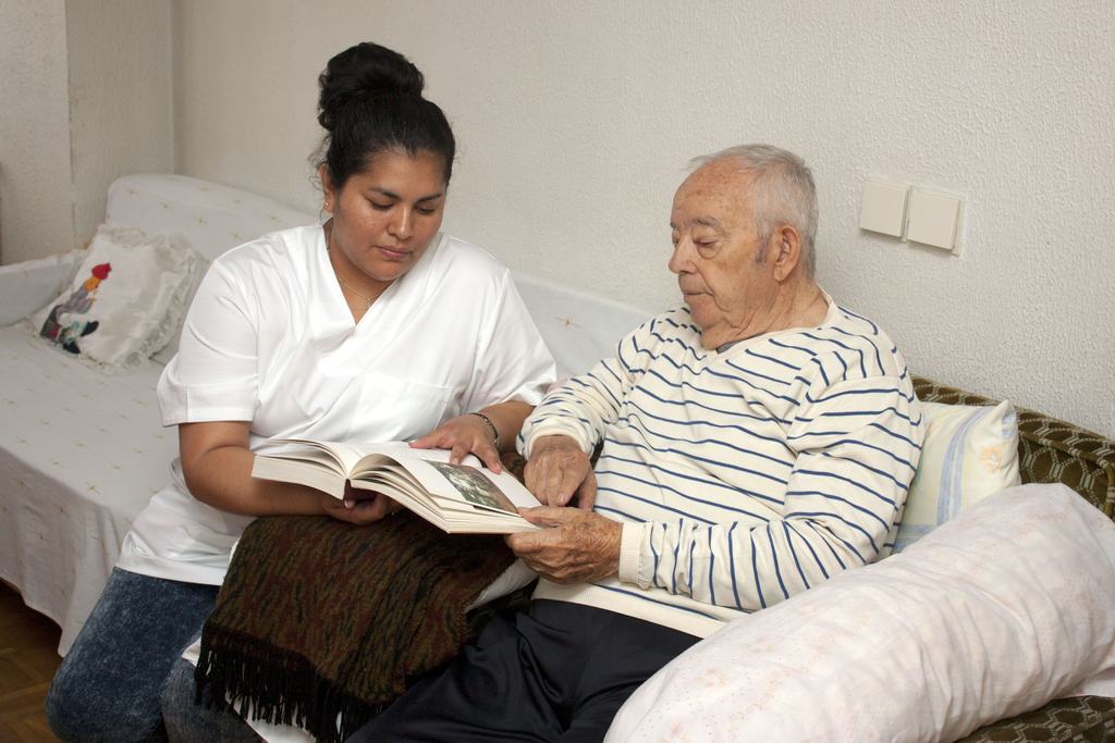 A care worker sitting beside an older man at home, reading through a book with him.